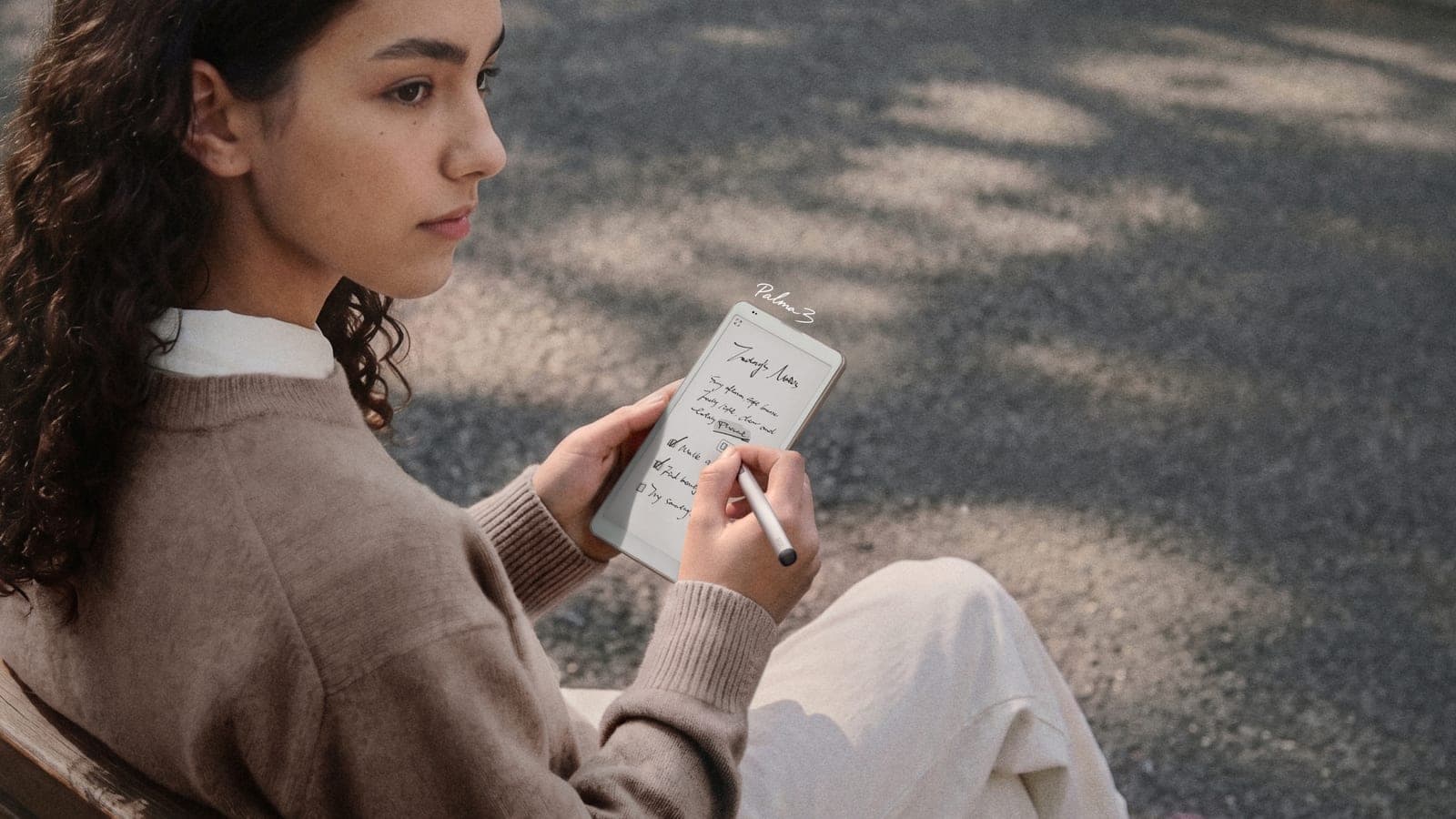 Une lectrice assise dehors écrit une liste de tâches sur un BOOX Palma 3 gris titane avec le stylet InkSense Plus